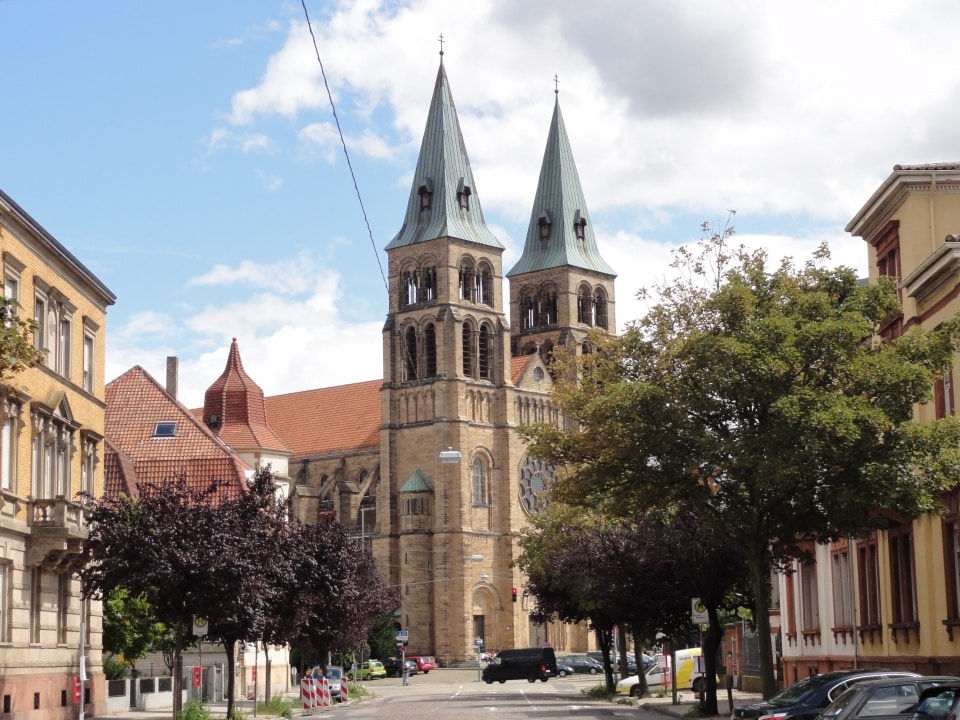 Zweiflügelige Steinkirche mit gewölbten Fenstern, von einer von Bäumen gesäumten Straße aus gesehen, bei teilweise bewölktem Himmel.