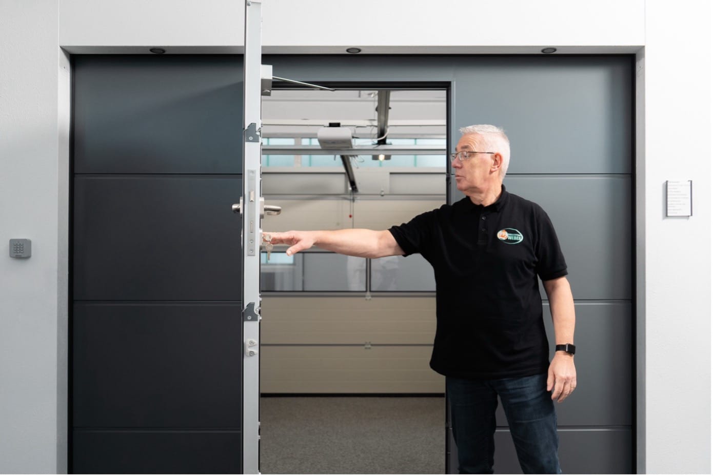 Mann im schwarzen Polohemd, der eine moderne graue Tür öffnet, in einem hellen Innenraum stehend.