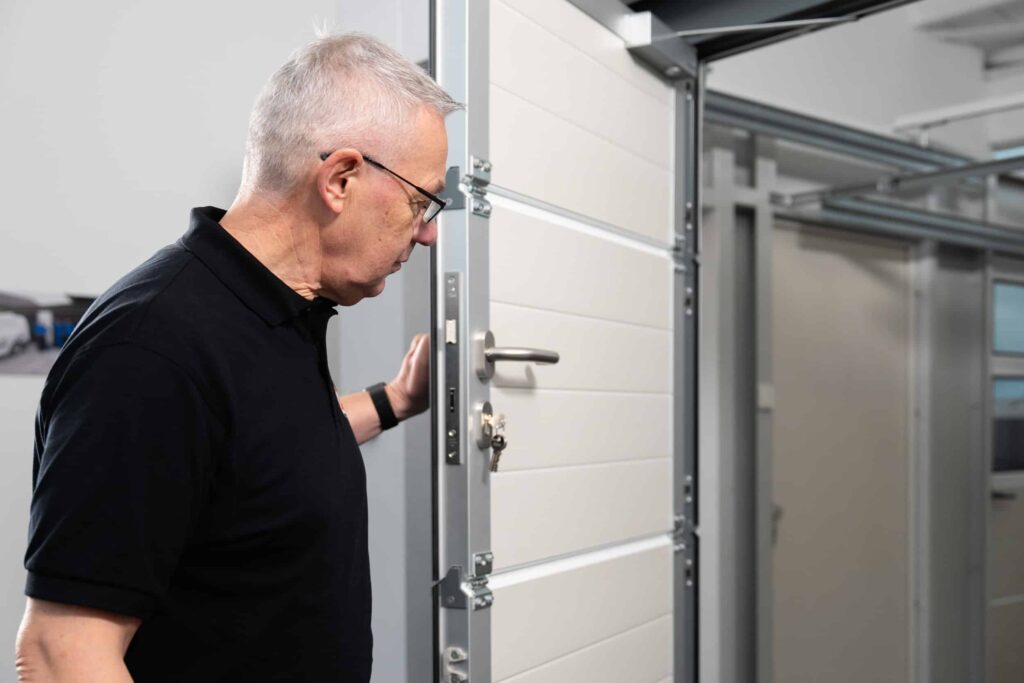 Älterer Mann mit Brille und schwarzem Hemd inspiziert ein teilweise geöffnetes weißes Garagentor in einem Haus.