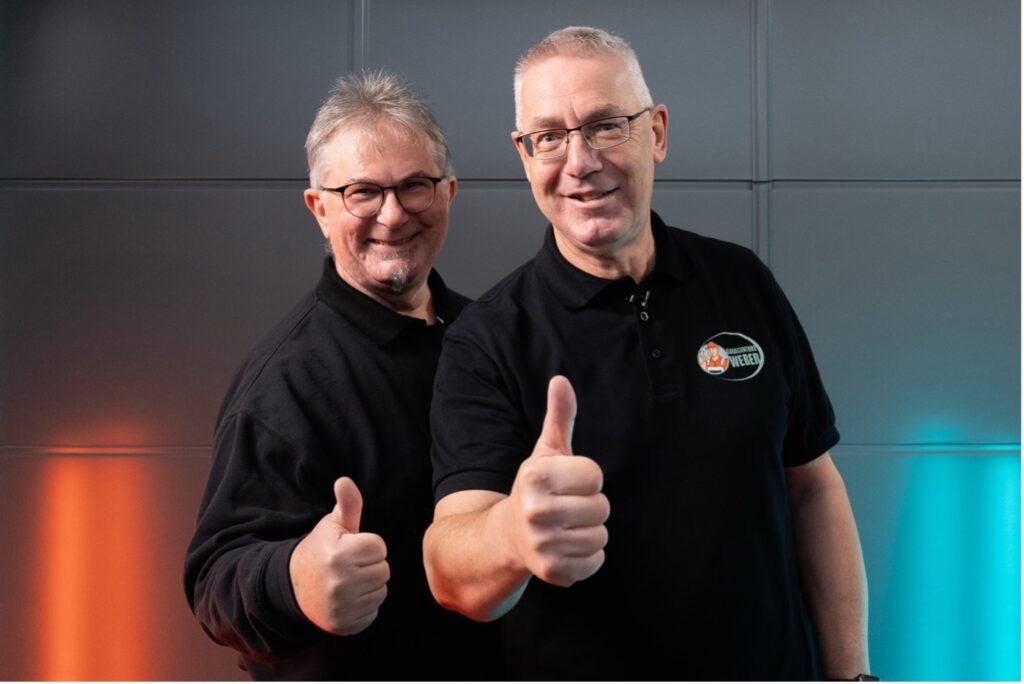 Zwei lächelnde Männer in schwarzen Hemden, die den Daumen nach oben strecken, stehen vor einer grauen Wand.