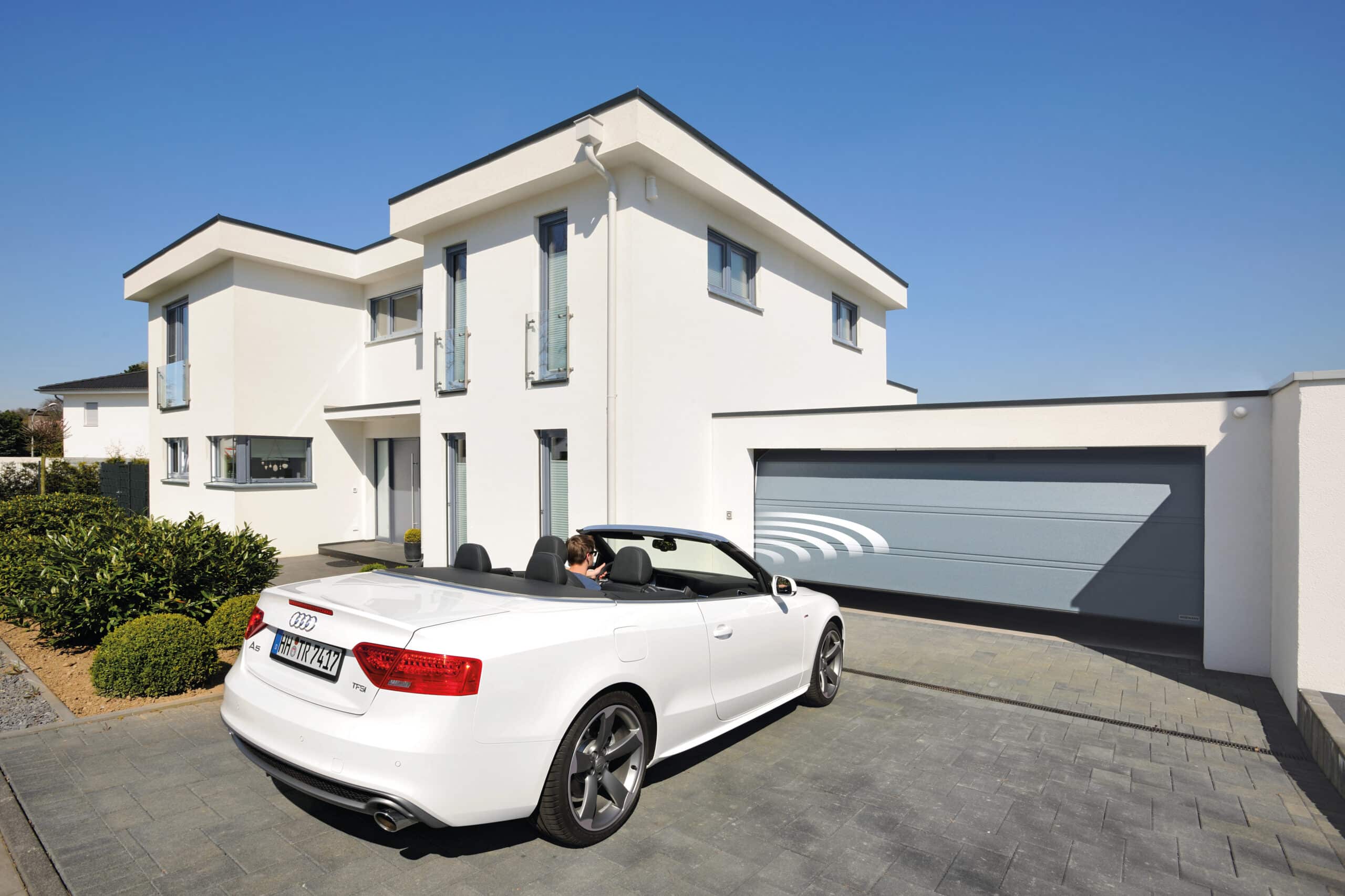 Ein weißes Cabriolet parkt vor einem modernen weißen Haus mit grauem Garagentor unter einem klaren Himmel.