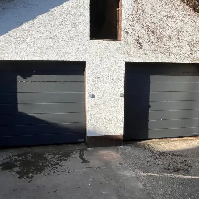 Zwei schwarze Garagentore unter einer weißen Wand mit einem offenen, türlosen Raum über den Garagen.