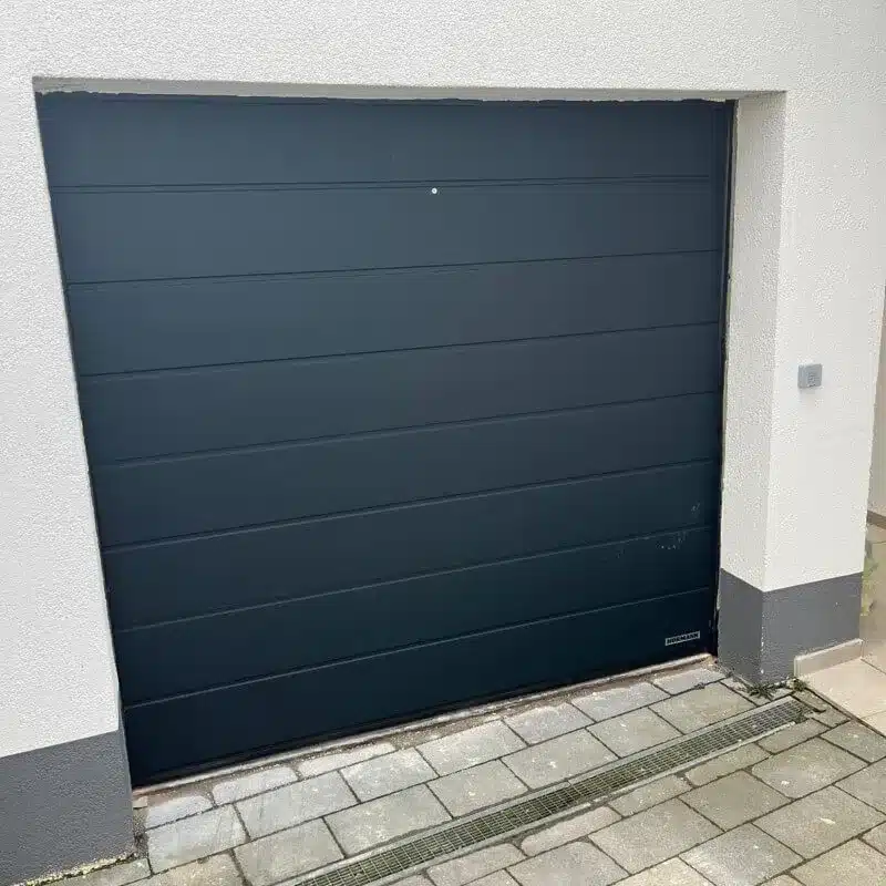 Ein schwarzes Garagentor, das schief eingebaut ist, mit ungleichen Spalten auf beiden Seiten, eingefasst in eine weiße Wand.