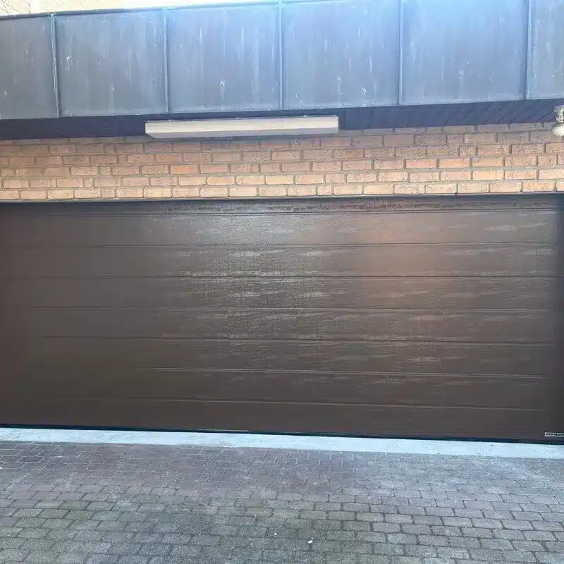 Ein geschlossenes dunkelbraunes Garagentor in einer Backsteinmauer mit einer darüber angebrachten Leuchte.