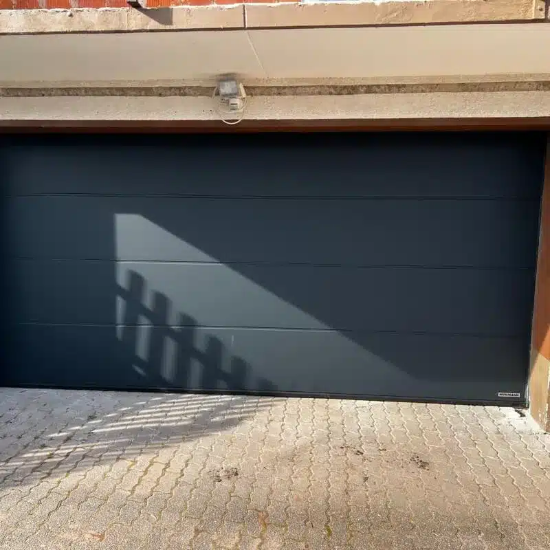 Geschlossenes dunkelgraues Garagentor, auf das das Sonnenlicht einen Zaunschatten wirft, und davor Pflastersteine.