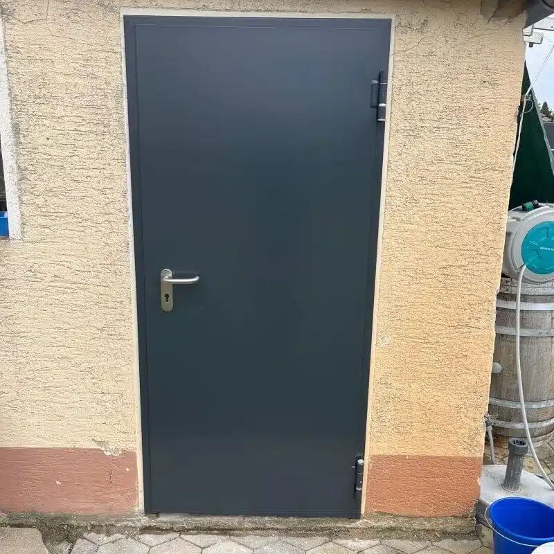 Eine geschlossene graue Metalltür in einer beigen Stuckwand mit einem blauen Eimer daneben auf dem Boden.