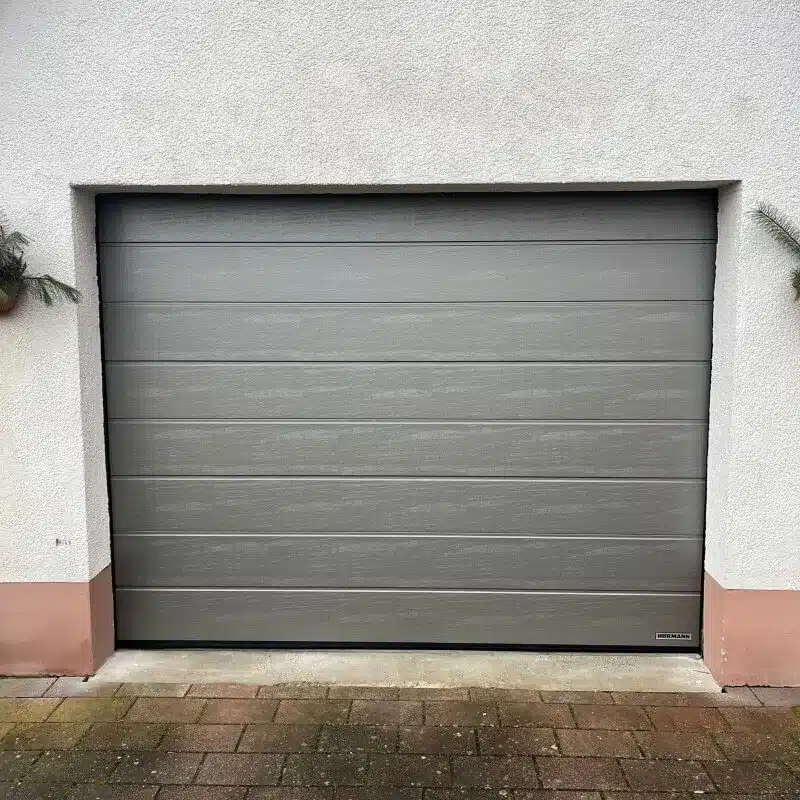 Ein geschlossenes graues Garagentor mit horizontalen Paneelen in einer weißen Wand über einer gemauerten Einfahrt.