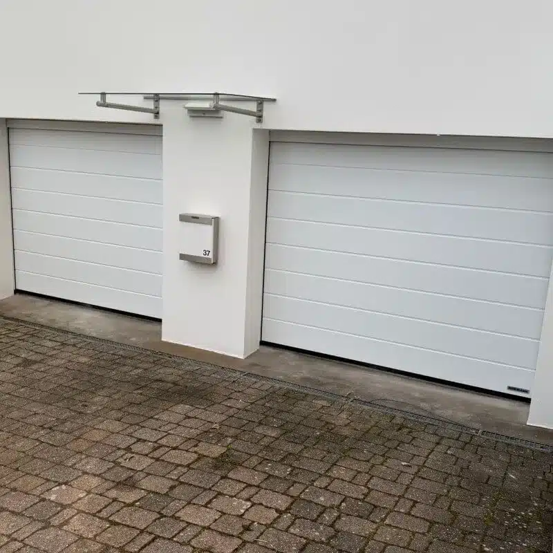 Zwei weiße Garagentore mit einem zentralen Briefkasten an einem modernen Gebäude über einer gepflasterten Einfahrt.