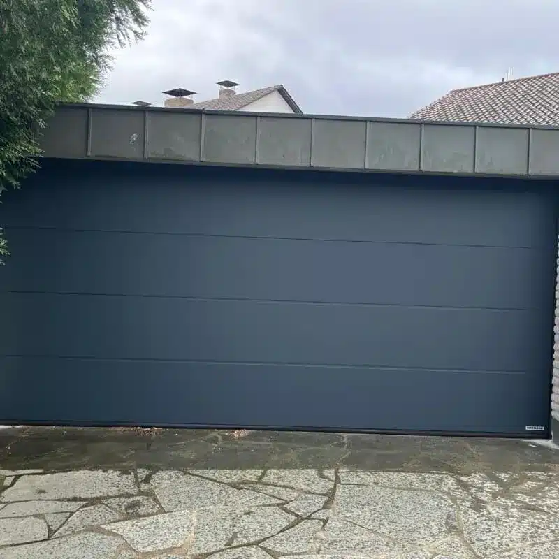 Ein modernes, dunkelgraues Garagentor mit einer Steinauffahrt und bedecktem Himmel.