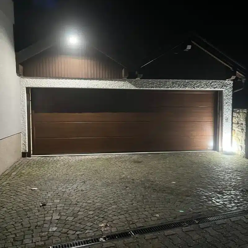 Ein großes hölzernes Garagentor bei Nacht, beleuchtet von hellen Außenlampen, mit einer Steinauffahrt davor.