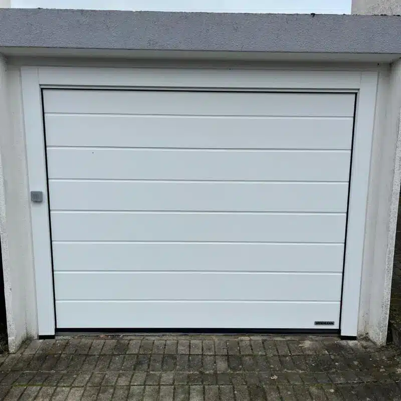 Geschlossenes weißes Garagentor mit horizontalen Paneelen in einem Betonrahmen über einer gepflasterten Einfahrt.